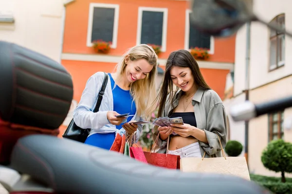 Two pretty excited young girls best friends, standing outdoors in city street with a lot of shopping bags and counting their money, satisfied after lucky shopping together — Stock Photo, Image