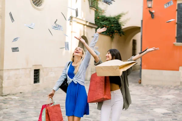 Two pretty young Caucasian women, wearing summer casual clothes, walking together with shopping bags and throwing paper money in the air. Shopping in the city, women friendship concept — Stock Photo, Image