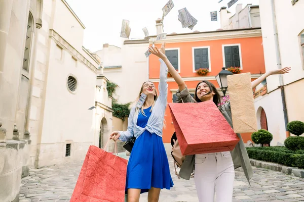 Two pretty young Caucasian women, wearing summer casual clothes, walking together with shopping bags and throwing paper money in the air. Shopping in the city, women friendship concept — Stock Photo, Image