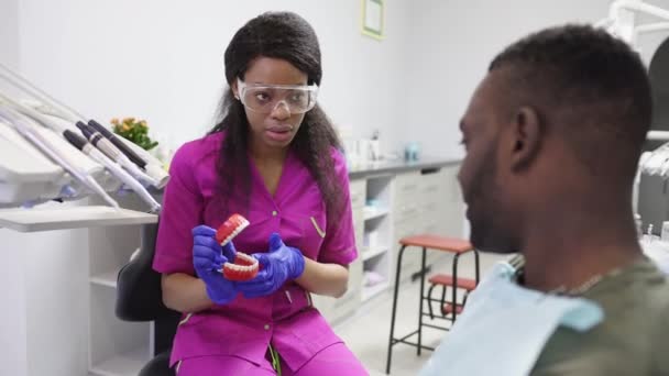 Güzel Afrikalı kadın dişçi erkek hastasıyla konuşuyor, yapay çene gösteriyor ve diş tedavisinin yollarını diş muayenehanesinde açıklıyor. Diş ve sağlık hizmetleri kavramı. — Stok video