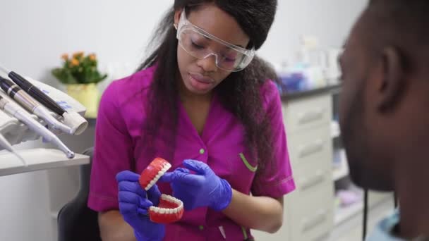 Primer plano de una joven dentista afroamericana sonriente que muestra un modelo de dientes a su paciente de piel oscura masculina en la clínica. Concepto de clínica dental . — Vídeos de Stock