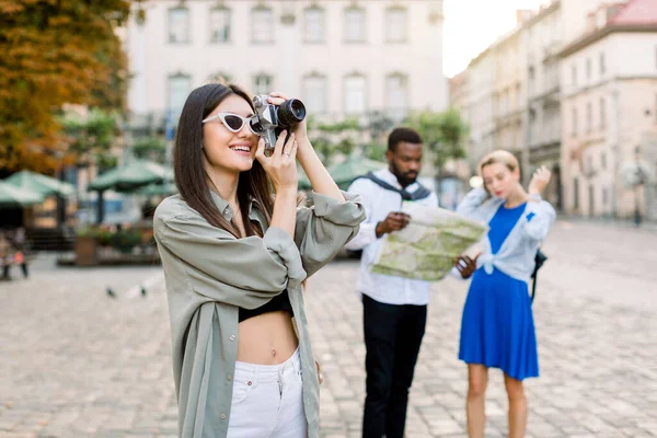 Mooie blanke brunette meisje toerist in casual kleding nemen van foto 's in de stad. Afrikaanse man en mooi blond meisje op zoek naar de stadsplattegrond voor een aantal interessante plaatsen, staan op de achtergrond — Stockfoto
