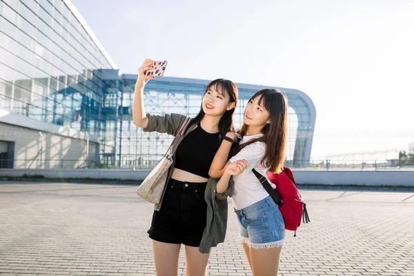 Concepto de turismo, amistad y viaje. dos alegre bien parecido jóvenes asiático niñas sonriendo y hacer selfie foto en el teléfono móvil, posando cerca de el moderno aeropuerto edificio antes de su viaje —  Fotos de Stock