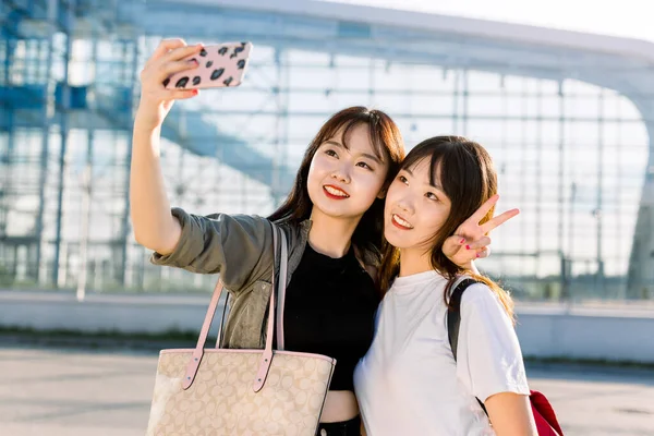 Dos encantadoras alegres sonrientes asiáticas mejores amigas chicas, abrazándose y mostrando gestos de señal de victoria durante la toma de foto selfie en el teléfono inteligente sobre el fondo del edificio del aeropuerto —  Fotos de Stock