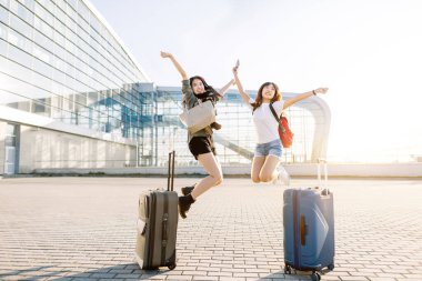 Seyahat ve tatil konsepti. Bavulları, pasaportları ve uçuş biletleri olan iki Asyalı genç kız havalimanı terminalinde birlikte atlayıp eğleniyorlar.