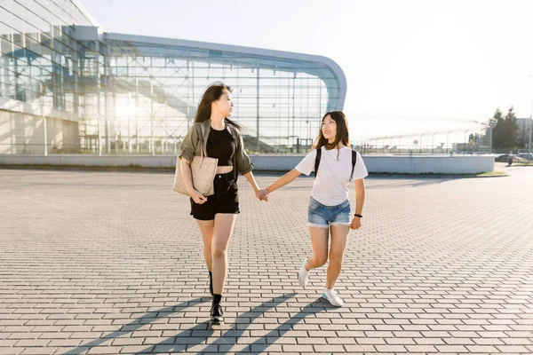 Turismo, aventuras y concepto de amistad. Retrato de verano de dos chicas asiáticas alegres, caminando juntas sobre el fondo del aeropuerto, cogidas de la mano y divirtiéndose antes de su viaje de vacaciones —  Fotos de Stock