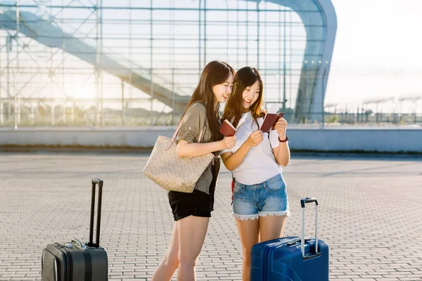 Dos hermosas mujeres asiáticas jóvenes felices con ropa casual, revisando sus pasaportes y boletos de avión, mientras están de pie cerca de la puerta del aeropuerto moderno con grandes maletas. Viaje conjunto, concepto de vacaciones —  Fotos de Stock