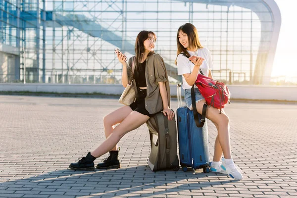 Літній портрет двох радісних азіатських жінок, що сидять на багажних валізах перед будівлею аеропорту і використовують свої телефони перед спільним польотом у відпустку — стокове фото