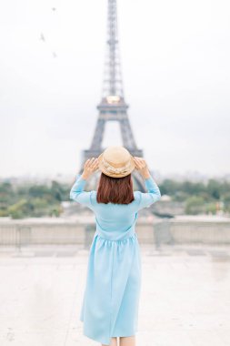 Şapkalı, mavi elbiseli, Paris 'teki Eyfel Kulesi' nin önünde poz veren çekici bir kadının arka plan portresi. Eiffel Kulesi 'nin manzarasının tadını çıkaran şık bir kadın. Seyahat, turizm konsepti