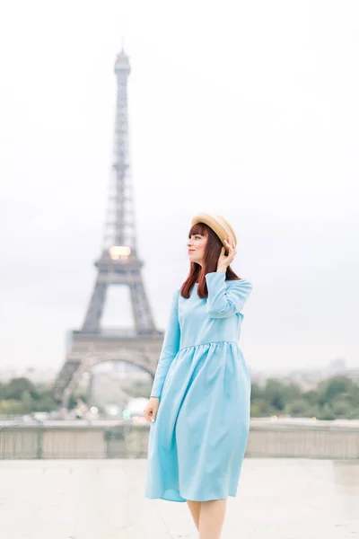 Romantická krásná dívka portrét s turné eiffel na pozadí. Pěkně jemná mladá dáma v modrých šatech a klobouku dotýkající se jejích zrzavých vlasů a chůze v Paříži — Stock fotografie
