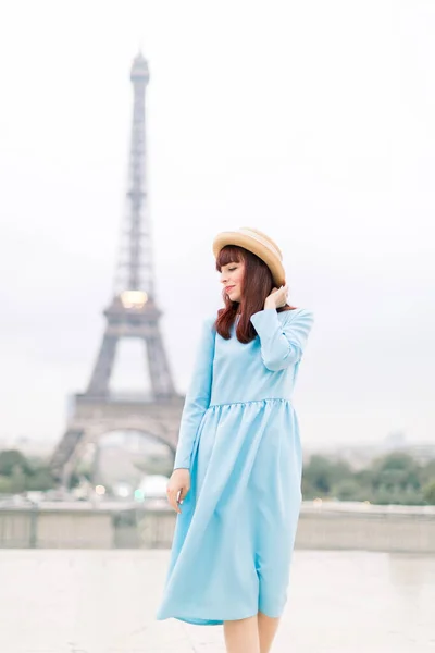 Krásná dívka na sobě dlouhé modré šaty a slamák klobouk, pózování a těší okamžik v Paříži s Eiffelovou věží na pozadí. — Stock fotografie
