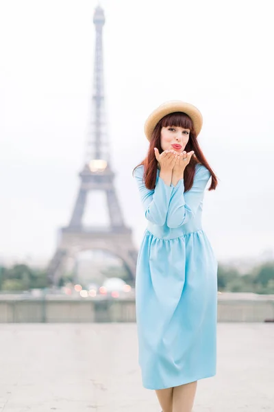 Ritratto di giovane donna elegante in cappello e abito blu che rende romantico il bacio dell'aria, mentre si trova sullo sfondo della torre Eiffel a Parigi — Foto Stock
