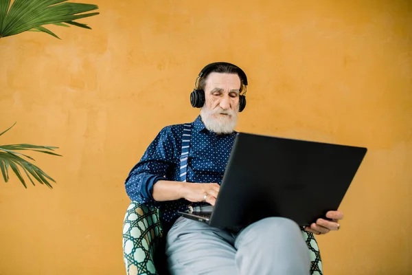 Estudio de tiro de hombre mayor concentrado guapo en ropa elegante, con auriculares, sentado en la silla y el uso de ordenador portátil. Personas mayores y tecnologías digitales —  Fotos de Stock