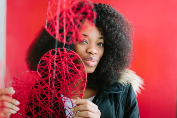 Ung vacker afroamerikansk flicka i svart rock ler på röd bakgrund med en dekorativ rött hjärta i händerna — Stockfoto
