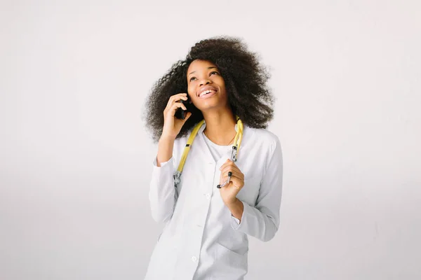 Portret van een jonge Afrikaanse arts in uniform met een stethoscoop in het ziekenhuis op witte achtergrond en telefonische gesprekken — Stockfoto