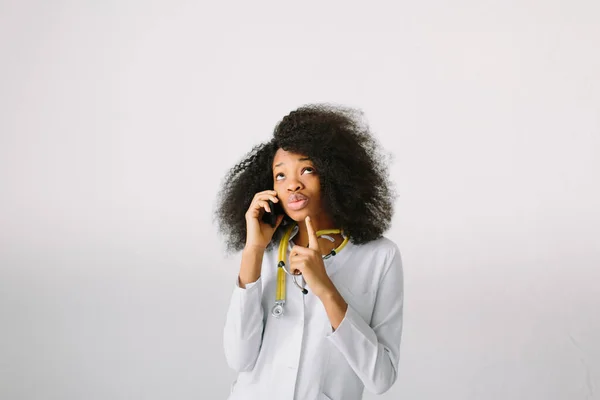 Ontwerp van reclame voor geneesmiddelen. Jong mooi meisje van Afro-Amerikaans meisje in witte jas met stethoscoop en map in het ziekenhuis. Een onpersoonlijke brede achtergrond. Praten aan de telefoon — Stockfoto
