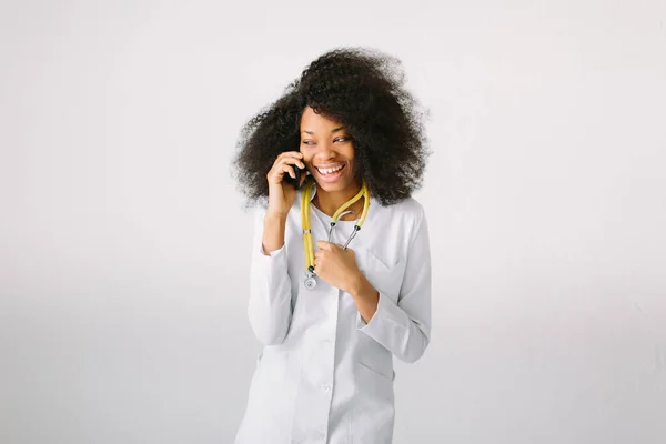 Ontwerp van reclame voor geneesmiddelen. Jong mooi meisje van Afro-Amerikaans meisje in witte jas met stethoscoop en map in het ziekenhuis. Een onpersoonlijke brede achtergrond. Praten aan de telefoon — Stockfoto