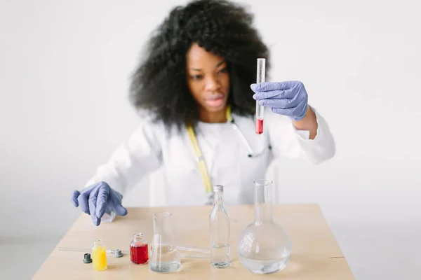 Joven hermosa chica afroamericana doctora en una bata blanca con un estetoscopio. sentado en una mesa con frascos de reactivo sobre fondo blanco. Mujer investigadora haciendo un análisis de sangre en un laboratorio — Foto de Stock