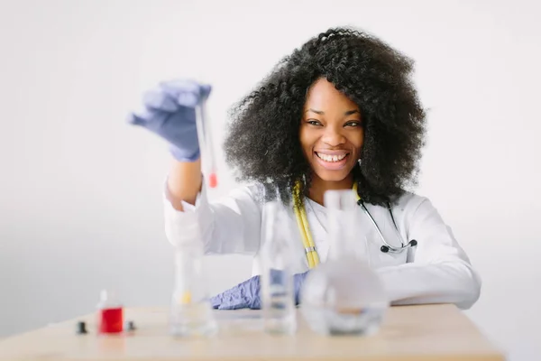 Young beautiful African American girl doctor in a white coat with a stethoscope. sitting at a table with reagent flasks on white background. Investigator checking test tubes. — Stock Photo, Image