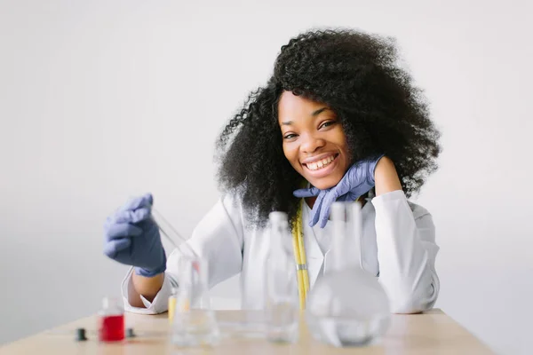 청진기가 달린 하얀 코트를 입은 아름다운 아프리카 계 미국인 소녀 의사. 하얀 배경에 에이전트 플래그와 테이블에 앉아 있습니다. 검사 관을 검사하는 수사관. — 스톡 사진