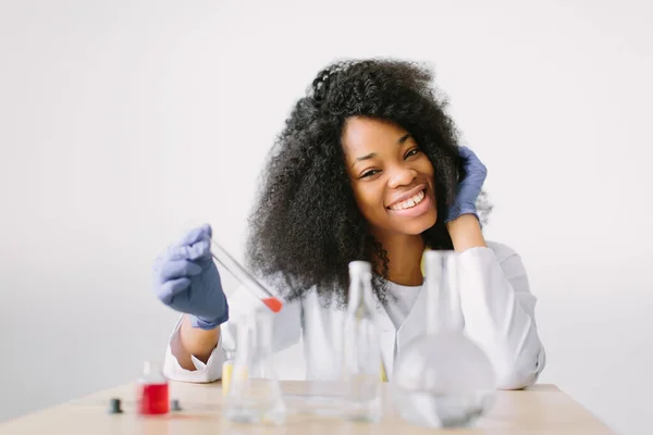 Młoda piękna Afroamerykanka lekarz w białym płaszczu ze stetoskopem. siedząc przy stole z kolbami odczynnika na białym tle. Badacz sprawdza probówki. — Zdjęcie stockowe