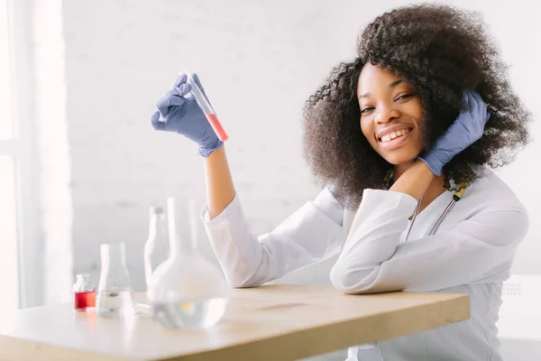 청진기가 달린 하얀 코트를 입은 아름다운 아프리카계 미국인 소녀 의사. 하얀 배경에 에이전트 플래그와 테이블에 앉아 있습니다. 분석을 위해 혈액 샘플을 연구하는 여성 연구원 — 스톡 사진