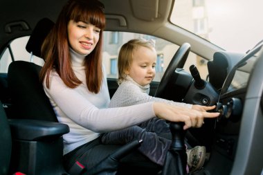 Arabada oynayan, müzik ya da radyo dinleyen, araba direksiyonunun yanındaki ön koltukta oturan küçük tatlı bir kız çocuğu olan genç bir anne. Aile gezisi, çocuk arabada.