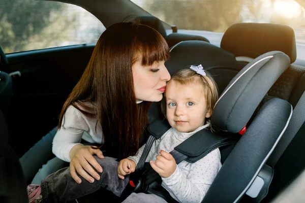 Retrato de uma jovem mãe ruiva bonita, sentada em um carro e beijando sua filhinha fofa em um assento de carro infantil. Viagem em família, conceito de segurança infantil — Fotografia de Stock