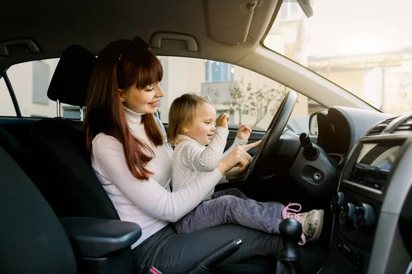 Mãe com criança pequena menina de joelhos mostra como dirigir um carro e o que é volante. Mãe e bebê se divertindo em um carro — Fotografia de Stock