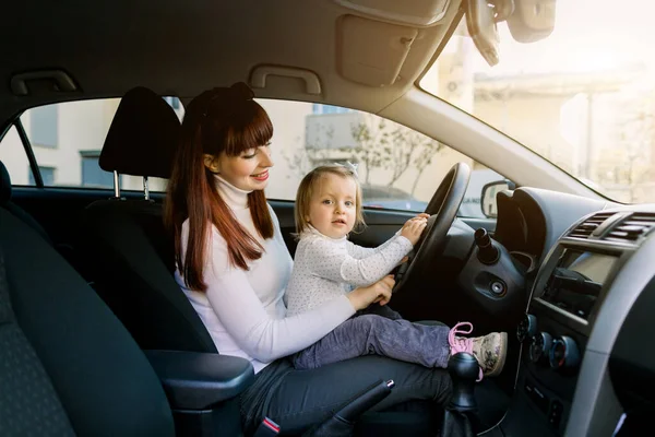 Jovem mãe com sua filhinha sentada no carro no banco da frente perto do volante do carro. Uma mulher e uma menina fingindo estar dirigindo. Mãe mostra seu bebê o carro dentro — Fotografia de Stock