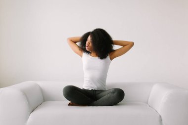 Beyaz yatakta lotus pozisyonunda oturan Afro-Amerikalı genç bir kadın.