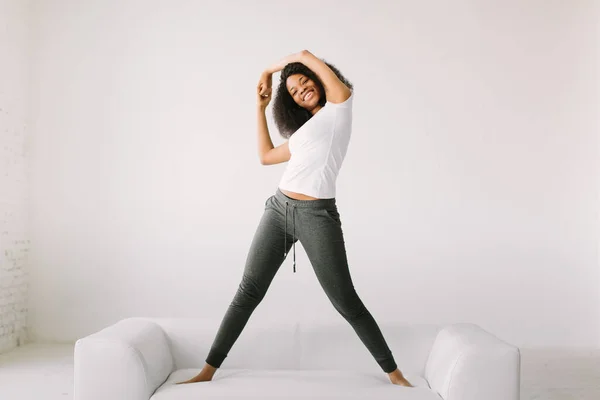 Eine junge Afroamerikanerin steht auf einem weißen Bett und macht körperliche Übungen — Stockfoto