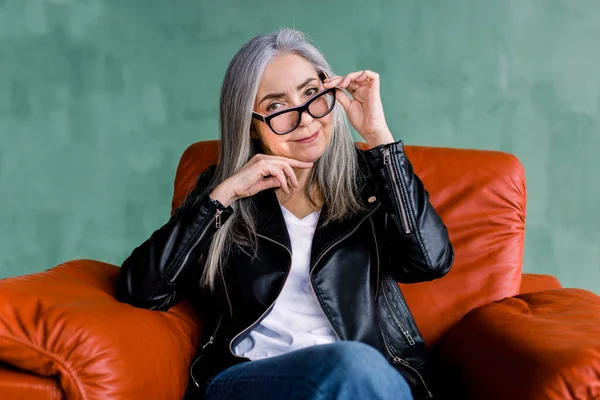 Pretty smiling 60-aged senior lady with long straight gray hair, looking at camera, while sitting in soft red armchair in front of green wall background and touching her eyeglasses — Stock Photo, Image