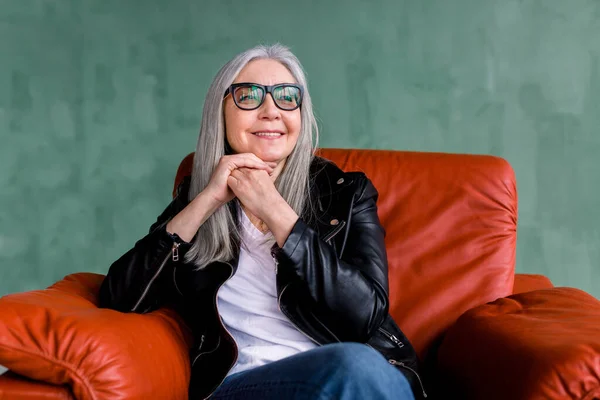Beautiful stylish senior woman with long straight gray hair, wearing eyeglasses and black leather jacket, sitting in comfortable sofr red armchair on green background and dreamly looking aside — Stock Photo, Image