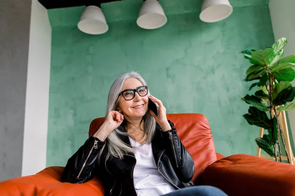 Bautiful joyful elderly woman with long straight gray hair, wearing leather jacket and eyeglasses, which talking with her friend by phone, sitting in red chair in stylish loft room with green wall — Stock Photo, Image