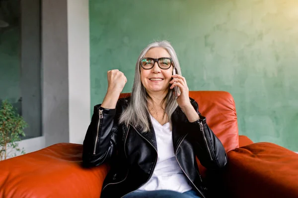 Attractive modern senior lady in black leather jacket, sitting in red soft armchair, keeping her fist clenched and smiling during mobile call with her best friend — Stock Photo, Image