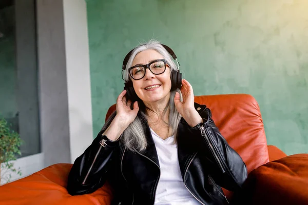 Charming joyful happy senior lady with long gray hair, wearing stylish black leather jacket, sitting in red armchair on green background and enjoying listening to music in earphones — Stock Photo, Image