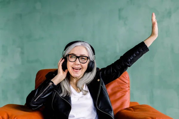 Happy positive pretty senior woman, dressed in stylish leather jacket, which enjoying favourite music in earphones and holding her arm up, while sitting in comfortable red armchair on green background — Stock Photo, Image