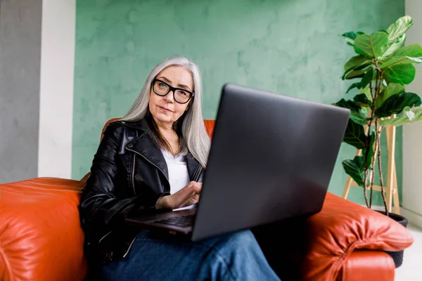 Πορτρέτο της ελκυστικής χαμογελαστή μοντέρνα ηλικιωμένη γυναίκα φορώντας γυαλιά ηλίου και μαύρο δερμάτινο μπουφάν, ποζάροντας στην κάμερα ενώ κάθεται σε άνετη κόκκινη πολυθρόνα και χρησιμοποιώντας τον φορητό υπολογιστή της — Φωτογραφία Αρχείου