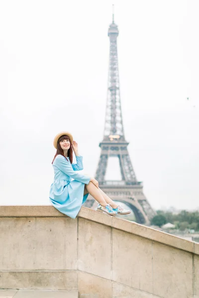 Belle fille rousse parisienne au chapeau élégant et robe bleue assise dans les escaliers avec le fond de la tour Eiffel, et levant les yeux avec un beau sourire — Photo