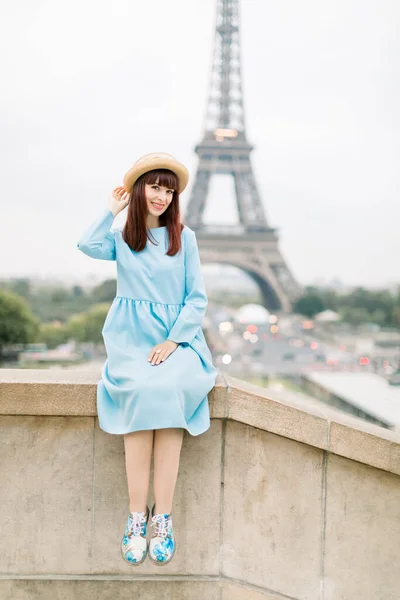 Paříž, Francie. Pěkně usměvavá žena turista v modrých šatech a klobouku, sedí na schodech pro fotografii před Eiffelovou věží, těší slavné turistické místa v Paříži — Stock fotografie