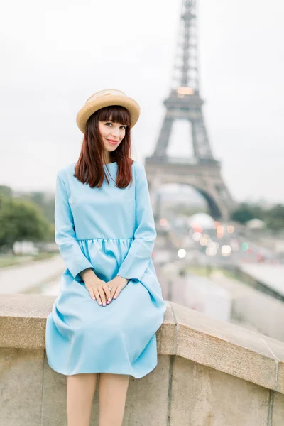 Venkovní obraz nádherné mladé ženy v klobouku a modré šaty sedí na schodech v krásném evropském městě. Paříž, Francie, Eiffelova věž na pozadí. Koncept cestování a osob — Stock fotografie
