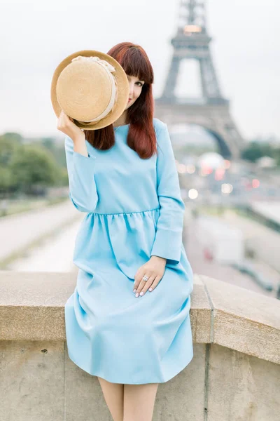 Giovane bella donna dai capelli rossi in abito blu, guardando la fotocamera, seduto sulle scale Trocadero e giocando con il cappello, nascondendo la faccia. Torre Eiffel sullo sfondo, Parigi, Francia . — Foto Stock