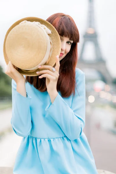 Giovane bella donna dai capelli rossi in abito blu, guardando la fotocamera, seduto sulle scale Trocadero e giocando con il cappello, nascondendo la faccia. Torre Eiffel sullo sfondo, Parigi, Francia . — Foto Stock