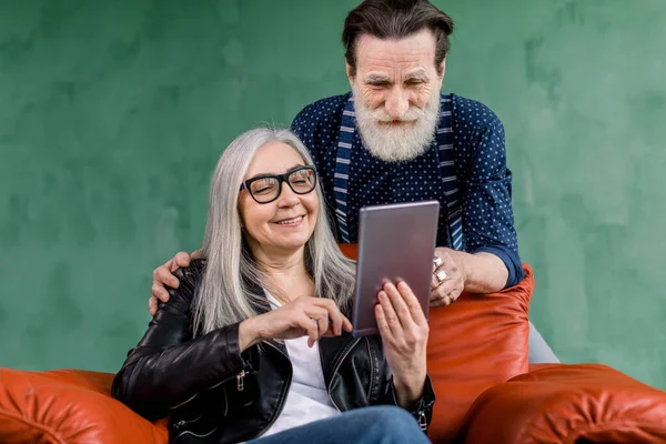 Close up indoor portrait of smiling attractive senior couple, bearded man and gray haired woman, using digital tablet for spending time together, browsing internet or social networks — Stock Photo, Image