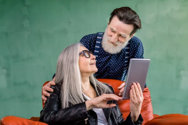 Στούντιο πλάνο του ευτυχισμένου ηλικιωμένου ζευγαριού της οικογένειας, κομψό άνδρα και γυναίκα, κοιτάζοντας ο ένας τον άλλον κατά την περιήγηση στο διαδίκτυο ή χρησιμοποιώντας εφαρμογές σε tablet i-pad και μιλάμε ο ένας τον άλλον. Η γυναίκα κάθεται στην κόκκινη καρέκλα. — Φωτογραφία Αρχείου
