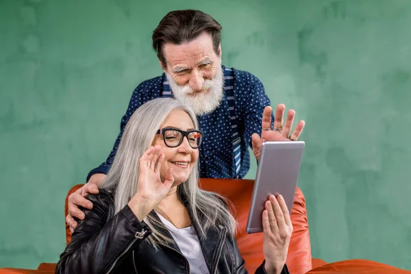 Αρκετά χαρούμενη 60-70 γκρίζα μαλλιά κυρία κάθεται σε μαλακή κόκκινη καρέκλα και κρατώντας tablet για βιντεοκλήση, ενώ όμορφος γενειοφόρος σύζυγος ακουμπά στην καρέκλα και την αγκαλιάζει, χαιρετώντας μαζί — Φωτογραφία Αρχείου