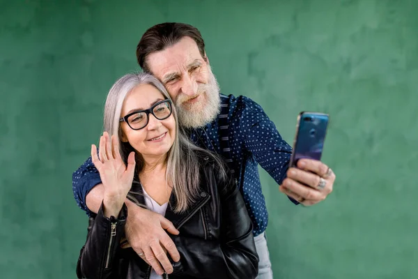 Πορτρέτο του ελκυστικού ηλικιωμένου ζευγαριού, στέκεται στο σπίτι μπροστά από το πράσινο τοίχο και μιλάει με τους φίλους τους μέσω τηλεφωνικής συνομιλίας βίντεο. Γενειοφόρος άντρας αγκαλιάζει τη γυναίκα του ενώ αυτή κουνάει το χέρι στην κάμερα. — Φωτογραφία Αρχείου