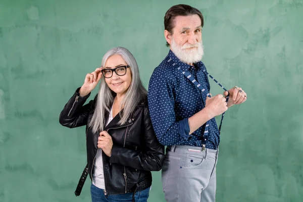 Lovely senior smiling joyful couple, handsome bearded man and pretty gray haired woman, posing to camera in front of green wall. Woman touching her eyeglasses, man pulling away his suspenders