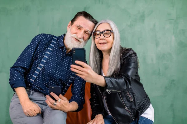 Attractive stylish couple, bearded man and gray haired lady, sitting together on the red chair and using the phone apps, looking at the smartphone screen and smiling. Isolated on green background. — Stock Photo, Image